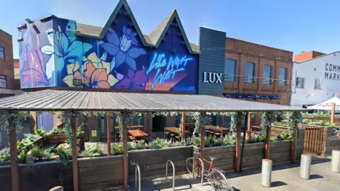 Google Maps The exterior of Lux nightclub in Belfast. It shows a colourful mural wall with 'life won't wait' in neon-style spray paint. There is also a sheltered beer garden.