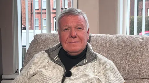 A man with short grey hair is pictured sitting in a living room in front of a large window. He is dressed in a black t-shirt with a light grey zip up jacket on top. He's sitting on a grey sofa. Behind him you can see red brick houses, a red car, and a white van outside the window. 