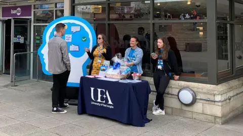 Students talk e the union bar. Three females are behind a table talking to a young man