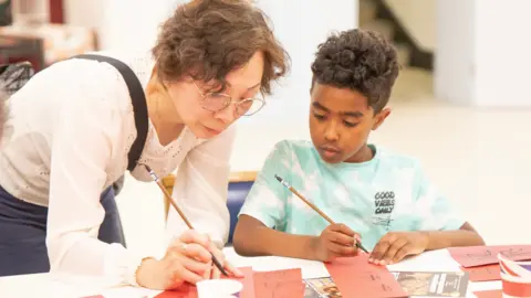 Shamphat A woman in a white top with glasses is teaching a boy in a blue top Chinese calligraphy. They are both using a brush and writing vertically from top to bottom on red card