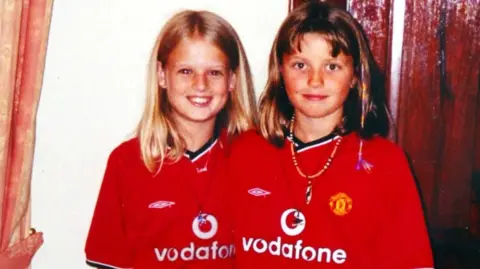 PA Media Two young girls dressed in matching red Manchester United shirts, smiling at the camera. 