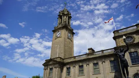The exterior of South Shields Town Hall with the clock tower in the centre of the image. It is an ornate, stone-built structure. An England flag is flying from another part of the building. The clockfaces sit on all four sides of the tower (only two are seen in the image) about halfway up. The tower itself has flat sides made of large stone until just above the clockfaces where it gives way to far more ornate masonry including statues and columns.