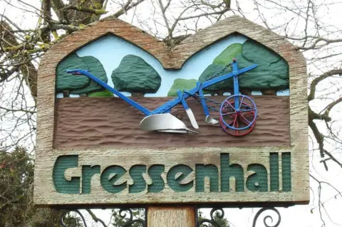 Geograph A carved village sign for Gressenhall featuring a blue horse-drawn plough mounted against a stylised countryside scene with green trees and tilled earth. The wooden sign is supported by decorative wrought-iron brackets, with leafless tree branches and village rooftops visible in the background.