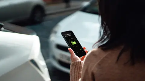 Getty Images A woman with long hair holds a mobile phone showing a generic taxi app, in front of two silver cars 