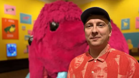 A man in a red shirt and cap smiles at the camera as he stands in a room with yellow walls, colourful paintings and a pink woolly mammoth sculpture behind him.