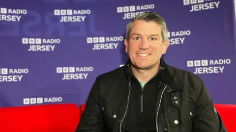 A picture of Dan McAlister Jersey RFC chairman staring at a camera. He has grey and black hair and is smiling. He is wearing a black quarter zip shirt with a black jacket on top.