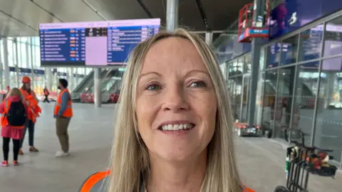 A blonde haired woman wearing an orange hi-vis vest stands in front of a large blue screening showing a list of times and destinations. There are a group of people wearing hi-vis vests in the background talking to each other.