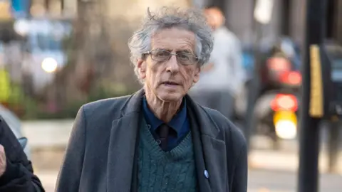 PA Media Piers Corbyn with grey hair and glasses walks towards the camera. He also wears a grey coat, grey trousers and a dark green jersey. 