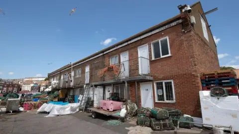North Yorkshire Council A harbour yard area with a two-storey brick building. Equipment associated with fishing activities is spread across the area, including stacks of lobster pots and crab traps. Coiled ropes and nets are laid on the ground, with white-wrapped pallets stacked to the right alongside loose rope offcuts and tools.