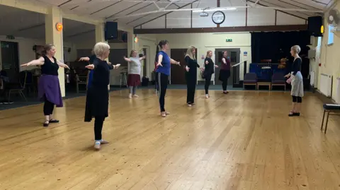 Aimee Deter/BBC Women are forming two rows and are standing in a village hall which has light brown wooden floors. They are all doing ballet and looking towards the instructor.