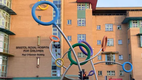 The entrance to Bristol Royal Hospital for Children. There is an abstract sculpture outside the hospital made of tall metal poles with colourful circles on the top.