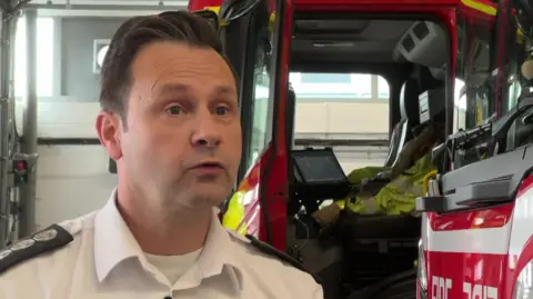 A man in a fire officer's white shirt and uniform. He is stood in front of the open door of a fire engine which is parked inside. 