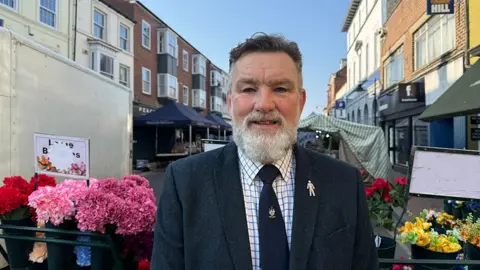 Grace McGrory / BBC Councillor Mark Blakeston he has shirt black hair and a grey beard and is wearing a black jacket, white checked shirt and black tie with a crest on it. He is standing in a street in Driffield surrounded by market stalls
