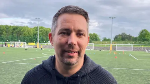 Chris Goreham/BBC Stevie Bramble is wearing a black hoodie in front of an astro turf football pitch, which has cones laid out on it. He has short dark hair and is smiling.