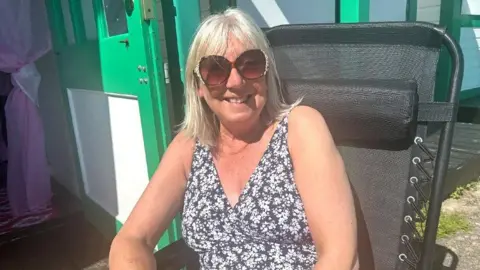 Liz Symons sits on a black, collapsible garden chair on the path outside her beach hut. She has very light-blonde bleached hair which is shoulder-length with a fringe. She is smiling, lightly-tanned, wears large sunglasses and a v-neck either sleeveless top or swimming costume which is purple-blue with a pattern of small white flowers. To her left one door of the hut is open showing a pink voile, see-through curtain which has been tied back in a knot half way down.