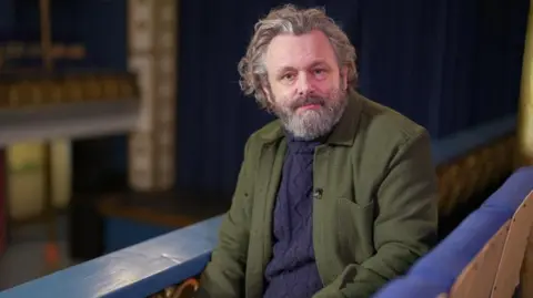 Michael Sheen is sitting in the gallery of a theatre, with the stage blurred out in the background. He is wearing a green jacket with a blue jumper. He has a grey beard and grey hair.