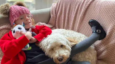 Holly Prince, photographed wearing a pink hat, a red cardigan and her grey school dress, smiles at the camera while sitting back on a sofa .She is cuddling a unicorn teddy while a fluffy cockapoo-type dog is resting on her torso. 