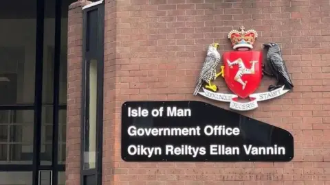 An Isle of Man Government sign, including a Manx coat of arms, on a red brick wall