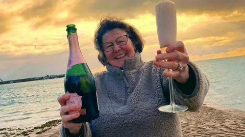 Rhona smiles at the camera as she holds a champagne glass and a bottle. She is wearing a grey zip up fleece and glasses. She has short brown hair. Behind her is a pier and the sun is setting as the sunshine breaks through the clouds.