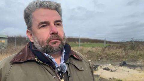 A man with salt and pepper hair and beard wearing a farmer's style coat looks off to the right 
