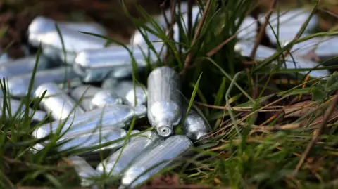 Discarded grey canisters of nitrous oxide, or laughing gas, lying in the grass. 