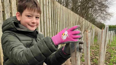 Mia Ambrose Isaac, 11, wears a green coat and pink gardening gloves as he grips a plastic tube which has a tree planter inside of it. He has brown hair and brown eyes. 