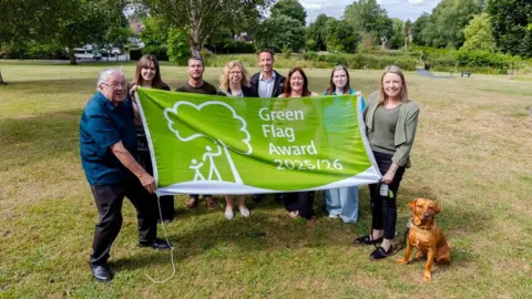 A group of men and women are standing in a green park with trees and bushes in the background. They are holding a large green and white flag between them that reads "green flag award 2025/26". There is a dog sitting next to the group, it is red in colour.