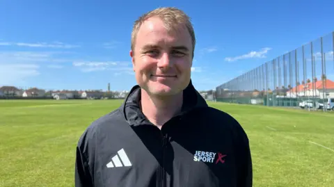 Dom is outside on a green playing field in the sunshine. He has a black jacket that says 'Jersey Sport' in white.