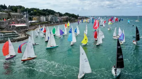 North Sails Cowes Week View of scores of yachts with sails up looking back towards the coast of Cowes
