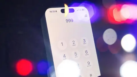 A mobile phone with 999 typed in with flashing lights in the background. 