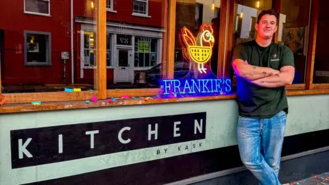 Adam leans against the restaurant windows, with his arms crossed and smiling at the camera. He wears a dark green t-shirt and blue jeans. 'Kitchen by Kask' is plastered on the outside of the restaurant, below the windows, and a 'Frankie's' sign lit up leans against the middle window pane. 