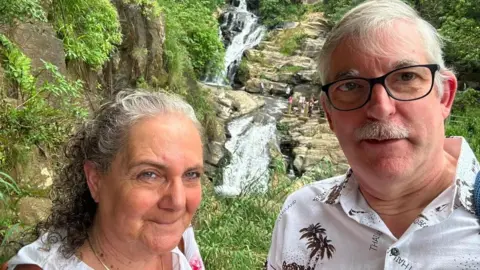 A couple in their 60s stand in front of a waterfall which has rocks and greenery around it. She is wearing a white shirt and gold chain necklace, her hair is shoulder length and clipped back from her face, and is grey and curly and she has the strap of a brown bag across her shoulder. He has white hair and a moustache and is wearing a white shirt with a palm tree pattern.