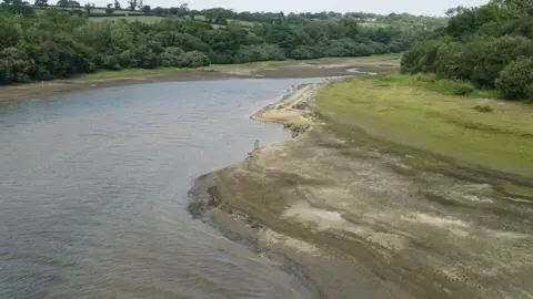 PA Media A view of low water levels at Roadford Lake in July 2022. The surrounding banks and trees are still green but the foreshore is brown and the water is too.