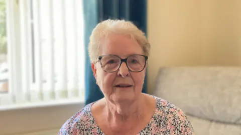 A woman with short hair is looking directly at the camera. She is wearing round dark framed glasses. She has a floral top on and is wearing a small chain necklace.