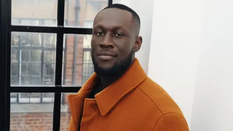 BBC Stormzy looking into the camera, with a slight smile on his face, standing to the side in front of a window, wearing a burnt orange coat. 