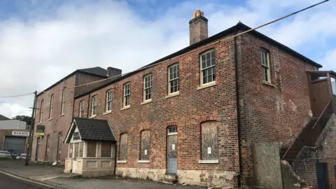 A boarded-up brick building of two storeys with a pitched roof and sash windows on the first floor.