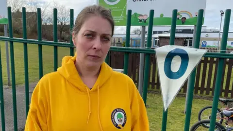 BBC/Seb Cheer Woman wearing a yellow hoodie looks into the camera. The hoodie has a logo for Queensway Primary School. She is standing in front of a fence, with a school in the background.