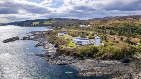 Sabhal Mòr Ostaig is a complex of white-walled buildings on a rocky coast. There is a landscape of trees and low hills behind the building.