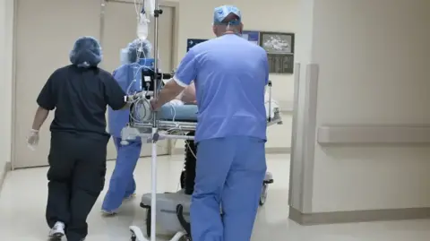 Getty Images Three members of NHS staff push a patient in a hospital bed along a corridor