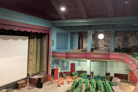 Supplied Abbeydale Picture House auditorium with a large white screen and blue painted walls with pink trim. There is a balcony. There are green seats and orange construction barriers as work progresses.