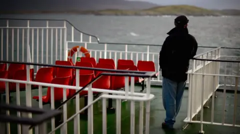 Getty Images A man wearing a black cap and jacket, blue jeans and black shoes stands on the deck of a CalMac ferry while he looks out to sea and the coastline beyond. There are white railings and red plastic seats on the deck.