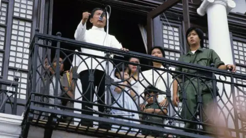 Getty Images Marcos Sr, wearing a white long-sleeved button-up shirt and black pants, raises his fist as he speaks into a microphone while standing on a balcony. Behind him is his family. Standing beside him is a young Bongbong Marcos, who is dressed in dark green overalls and putting one hand on the balcony railing.