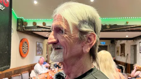 A side profile of Rick smiling with short hair after his hair was cut off for charity. A few customers in a pub can be seen in the distance.