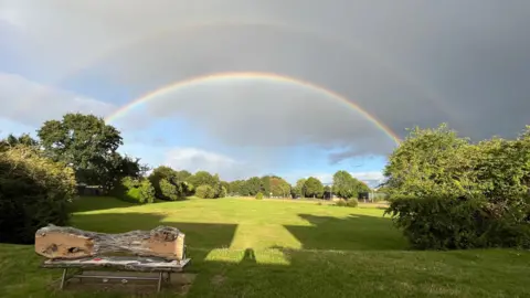 Two rainbows arch over a large green lawn in Thatcham, in front of a mixture of dark grey cloud and blue sky