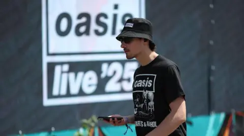 Adam Vaughan/EPA/Shutterstock An Oasis fan walks past advertising boards ahead of the first gig of the Oasis Live 25 tour in Cardiff.