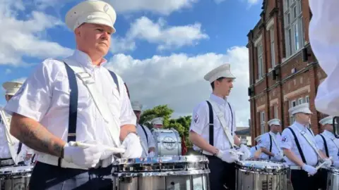BBC A group of men in white shirts and white gloves carry drums.