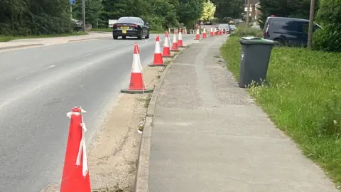 Graham Statter Line of red cones along a village street, with sporadic parked cars and a wheelie bin in view