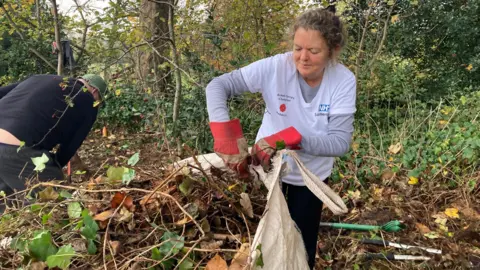 Rebecca Oliver wearing a light blue NHS Somerset T-shirt and red gardening gloves in a lightly wooded area. She is filling a white sack with overgrown plants that have been cleared. Another volunteer is also visible in the background, kneeling down and working at clearing the area.