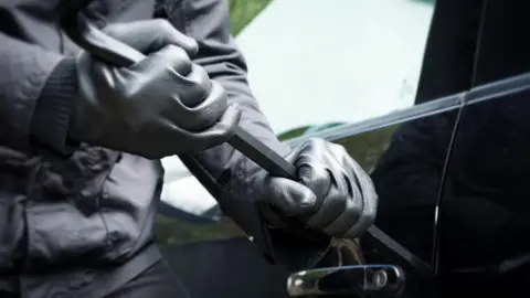 A man wearing black gloves using a crow bar to break into a car