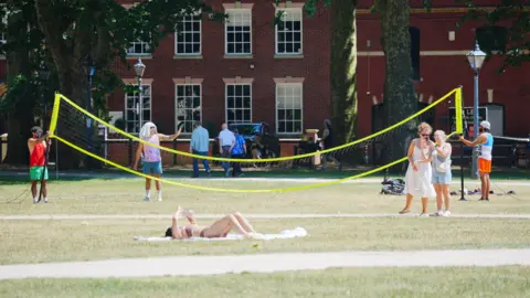 Ben Birchall/PA Wire A group of people wearing colourful vests and shorts are putting up a neon yellow volleyball net. Also in the picture is a young girl wearing a two-piece swim suit lying down on the grass and reading a book. There are also two women who have stopped to look at their phone.